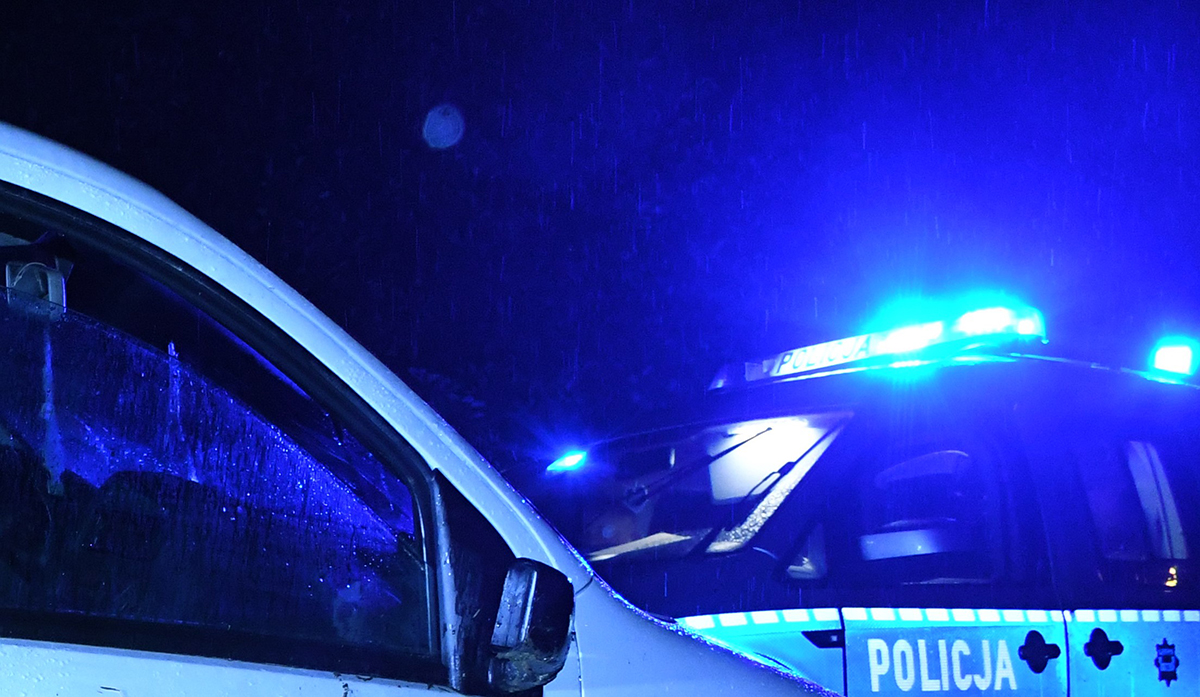 widoczny fragment samochodu białego po lewej stronie, w tle radiowóz policyjny, środek nocy, całe zdjęcie jest rozjaśnione tylko na niebiesko