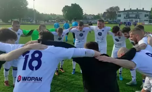 Piłkarze na murawie, stoją w kręgu trzymając się za ramiona, lekko pochyleni w kierunku środka kręgu
