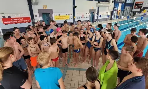 Młodzież w strojach pływackich (kilkadziesiąt osób, dziewczęta i chłopcy), stojąca w kółku. W tle tory pływackie na basanie pływalni zamkniętej