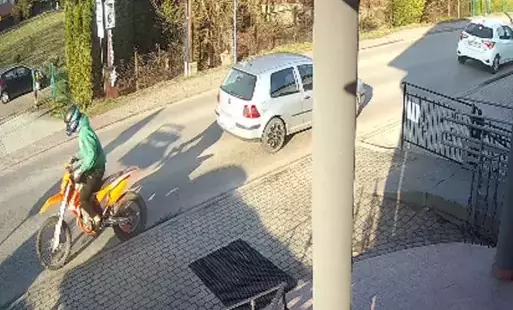 mężczyzna w zielonej bluzie z kapturem i czarnych spodniach siedzi na pomarańczowym motocyklu marki cross, przejeżdżając przez ulicę