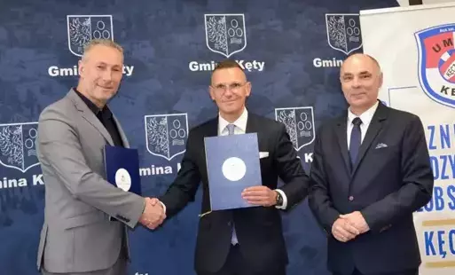 na niebieskim tle z białym godłem Gminy Kęty stoi grupa trzech mężczyzn - prezes GVM Carint oraz prezes UMKS Kęczanin Kęty ściskają sobie dłonie, po prawej stronie na tle białego baneru UMKS Kęczanin stoi I zastępca burmistrza Rafał Ficoń