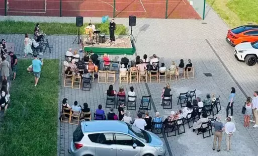 widok z okna na szkolny parking na którym odbywa się koncert, na podium stoi dwoje wykonawców, reszta osób w tle ogląda wydarzenie 