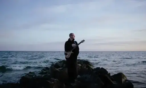 Mężczyzna z gitarą przewiedzona na pasku przez ramię stojący na skałach, w tle morze