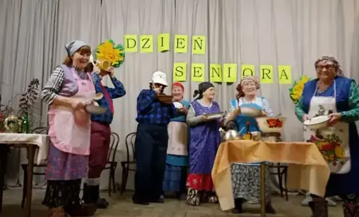 seniorzy w strojach scenicznych przygotowanych do przedstawienia stoją na scenie, za nimi na szarym tle jest napis dzień seniora