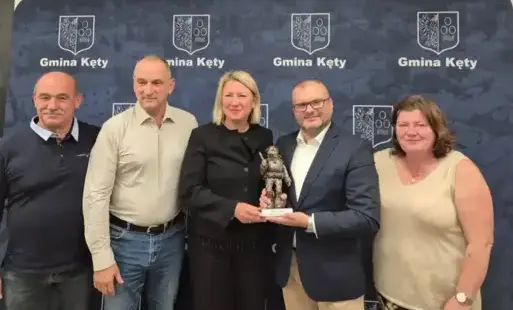 Pięcioro osób na tle ścianki promocyjnej z hermeb Gminy Kęty, konieta i mężczyzna stojący pośrodku prezentują statuetkę
