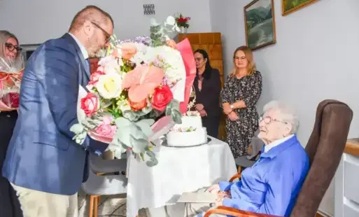 burmistrz gminy kęty marcin śliwa wręcza bukiet kwiatów solenizantce - pani Klementynie, siedzącej w fotelu po prawej stronie