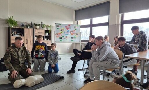 uczniowie siedzą w klasie, po lewej stronie kadru klęczy mężczyzna w mundurze przed manekinem szkoleniowym, wyjaśniając techniki udzielania pierwszej pomocy