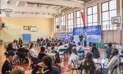 uczniowie siedzą przy stolikach w sali gimnastycznej, w tle bladoniebieski baner, przemawia wiceburmistrz gminy Kęty