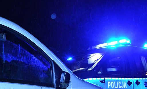 widoczny fragment samochodu białego po lewej stronie, w tle radiowóz policyjny, środek nocy, całe zdjęcie jest rozjaśnione tylko na niebiesko