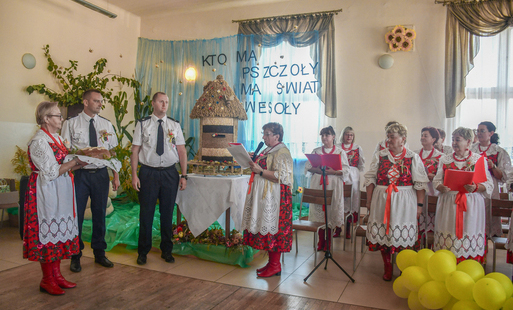po lewej stronie stoi gospodyni KGW Bielany trzymając chleb, w środku stoją prezes osp andrzej dziubek oraz naczelnik osp sławomir wójcicki, po prawej przewodnicząca KGW z mikrofonem, jest to moment wręczenia chleba gospodarzom uroczystości.