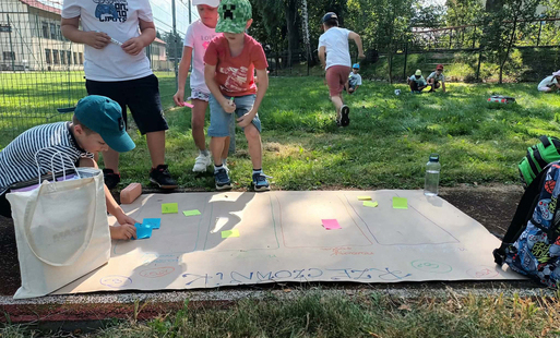 grupa dzieci uczęszcza na zajęcia szkolne na świeżym powietrzu - dzieci stoją nad planszą z tektury z napisem "rzeczownik" na podwórku