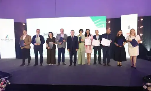 11 osób - kobiet i mężczyzn - stojących obok siebie, trzymających teczki na dokumnety. W tle ekran z napisami 